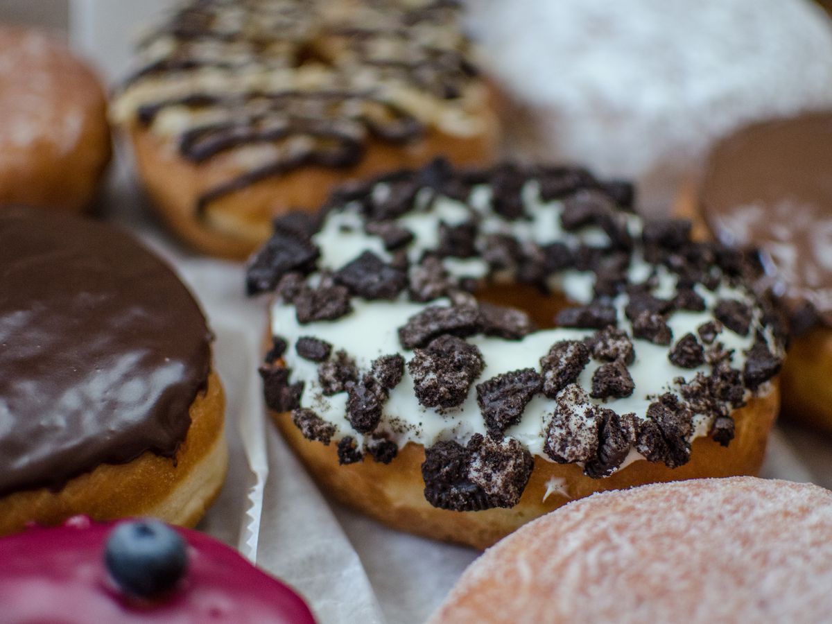 Closeup on several doughnuts on wax paper. The main doughnut in focus is topped with white frosting and Oreo cookie crumbles.