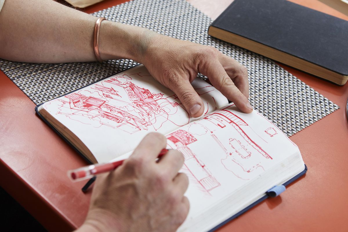 A sketchbook is being drawn in by a person. The hands and a red pencil are visible. The sketch is in red. There is a patterned mat underneath the open sketchbook. The table is red. 