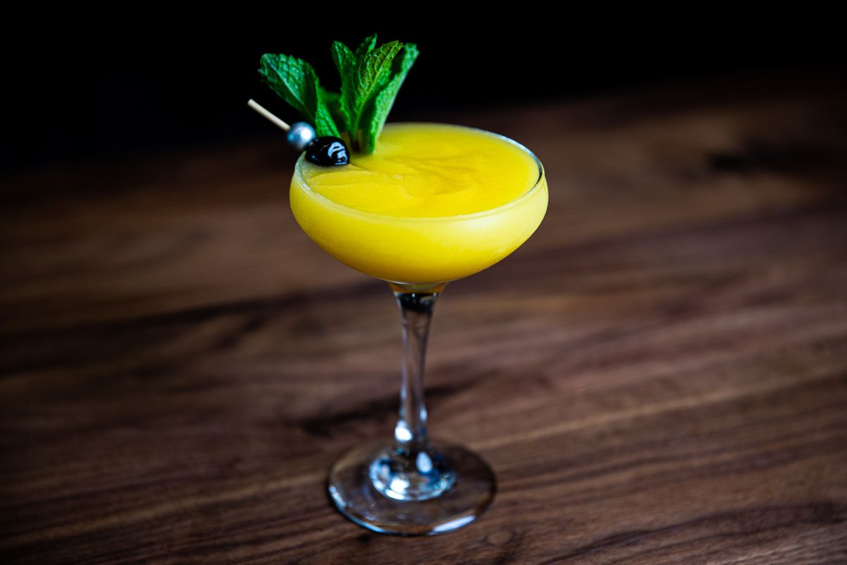 An orange cocktail in a coupe glass garnished with fresh mint.