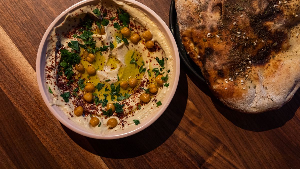 The hummus dip is served in a tan bowl with chopped cilantro and red sumac with olive oil and fried whole chickpeas on top. It’s served next to brown whole wheat flatbread with a spice mix on top.