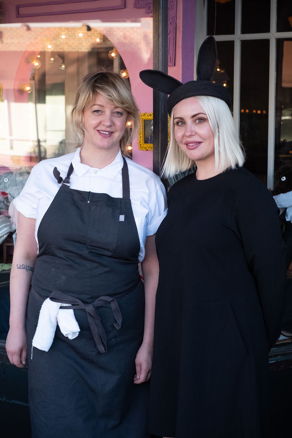 Chef Jamie Malone and Nikki Klocker outside Grand Cafe. Jamie wears an apron with a white towel tucked at the waist. Klocker wears a black sheath dress and a rabbit-eared chic hat