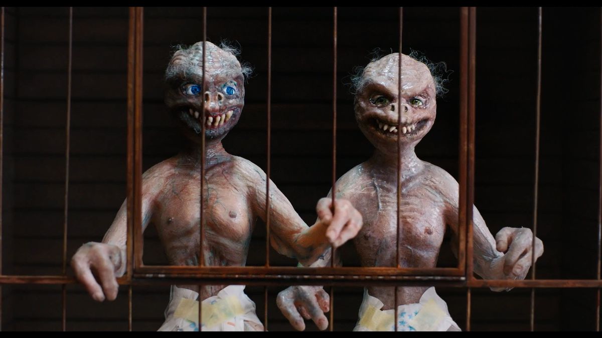 The hideous “Sewer Boys” puppets, two grinning, fanged, grey-skinned, slightly hairy little humanoid puppets in diapers, gripping the bars of their cage, in Dicks: The Musical