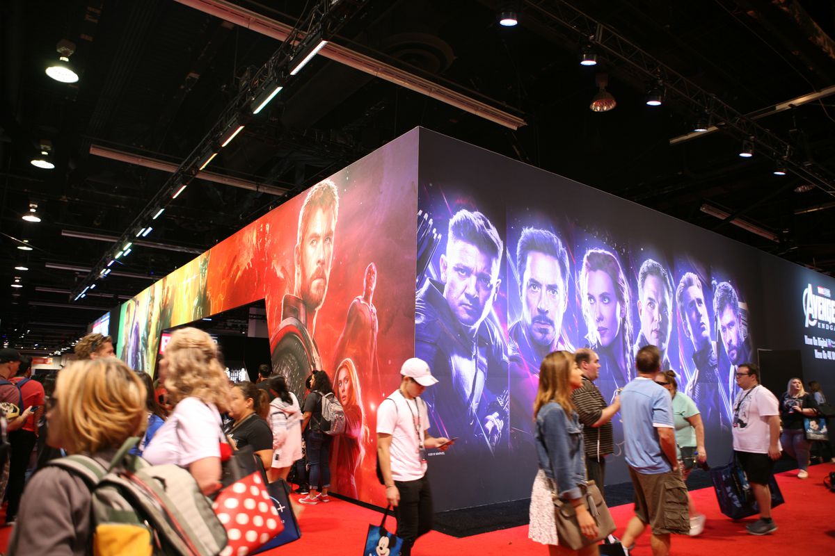 Fans at an expo walk past a huge display of Avengers superhero portraits.