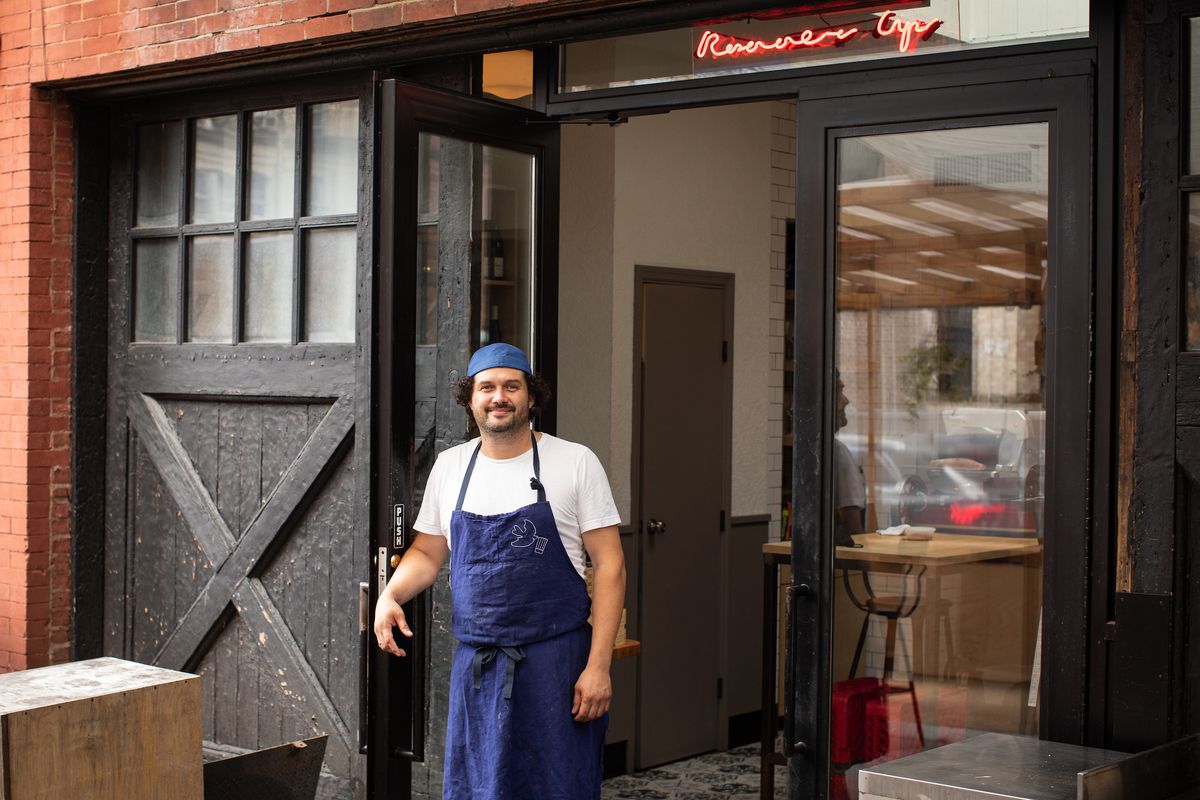 A man wearing a backwards hat and blue apron stands in front of a restaurant with floor-to-ceiling windows and a neon sign that reads “Runner Up”