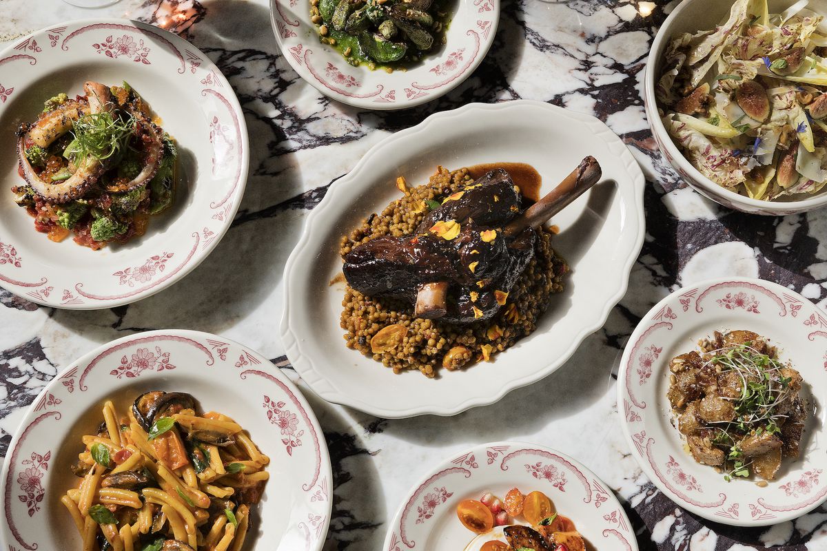 Overhead shot of a full marble table with octopus, salad, lamb shank with bone in, and more.