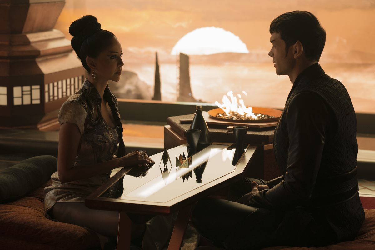 Gia Sandhu as T’Pring and Ethan Peck as Spock sit on cushions on either side of a small low table in a restaurant on Vulcan in Star Trek: Strange New Worlds. 