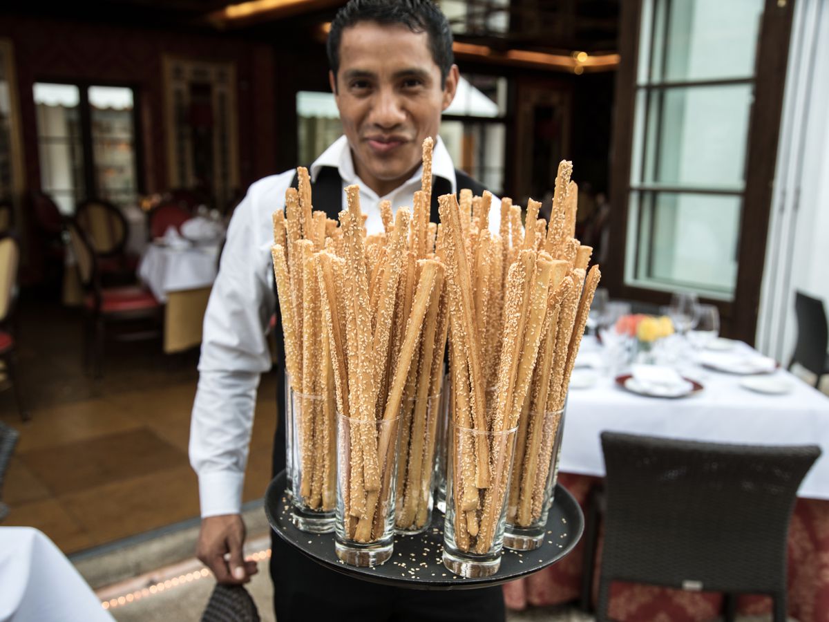 A waiter holds a tray full of grissini, Italian breadsticks. He’s looking directly at the camera. The courtyard of the restaurant is in the background and beyond the courtyard is the open doors of the inside of the restaurant.