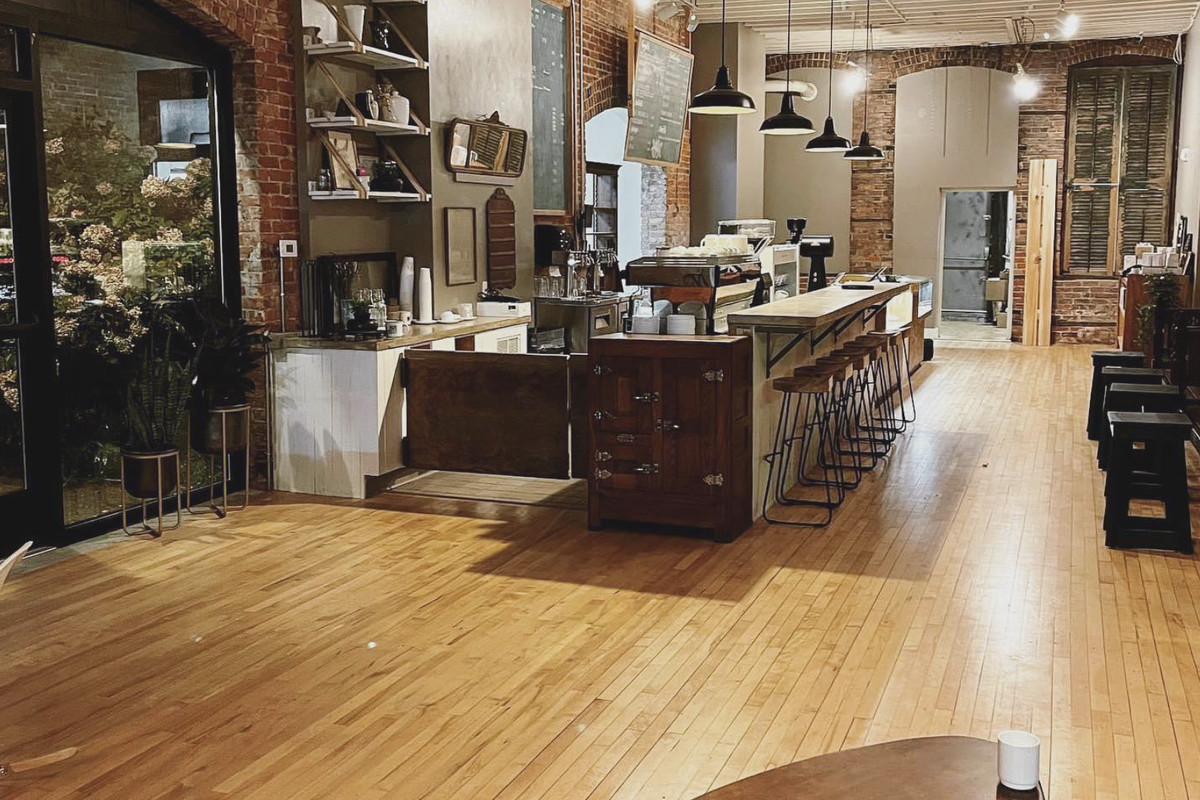 A coffee shop shows wooden floors and a counter with chairs for seating. There’s a large mirror against the exposed brick walls.
