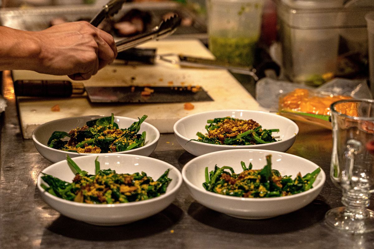 Four white bowls with green vegetables laid out on a kitchen table with hands in the background working over a cutting board