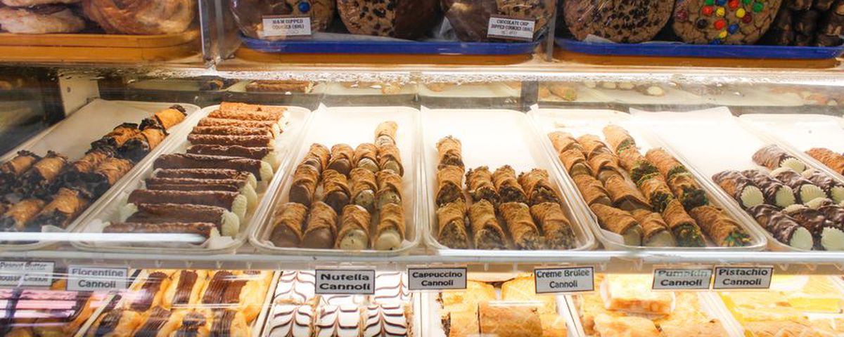 View of a pastry case in an old-fashioned Italian bakery, with rows of cannoli and other treats behind glass.