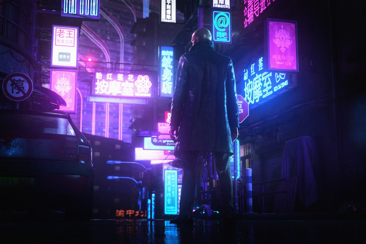 Agent 47 wearing a black trenchcoat standing on a rainy neon-lit street in Hitman 3’s Chongqing map