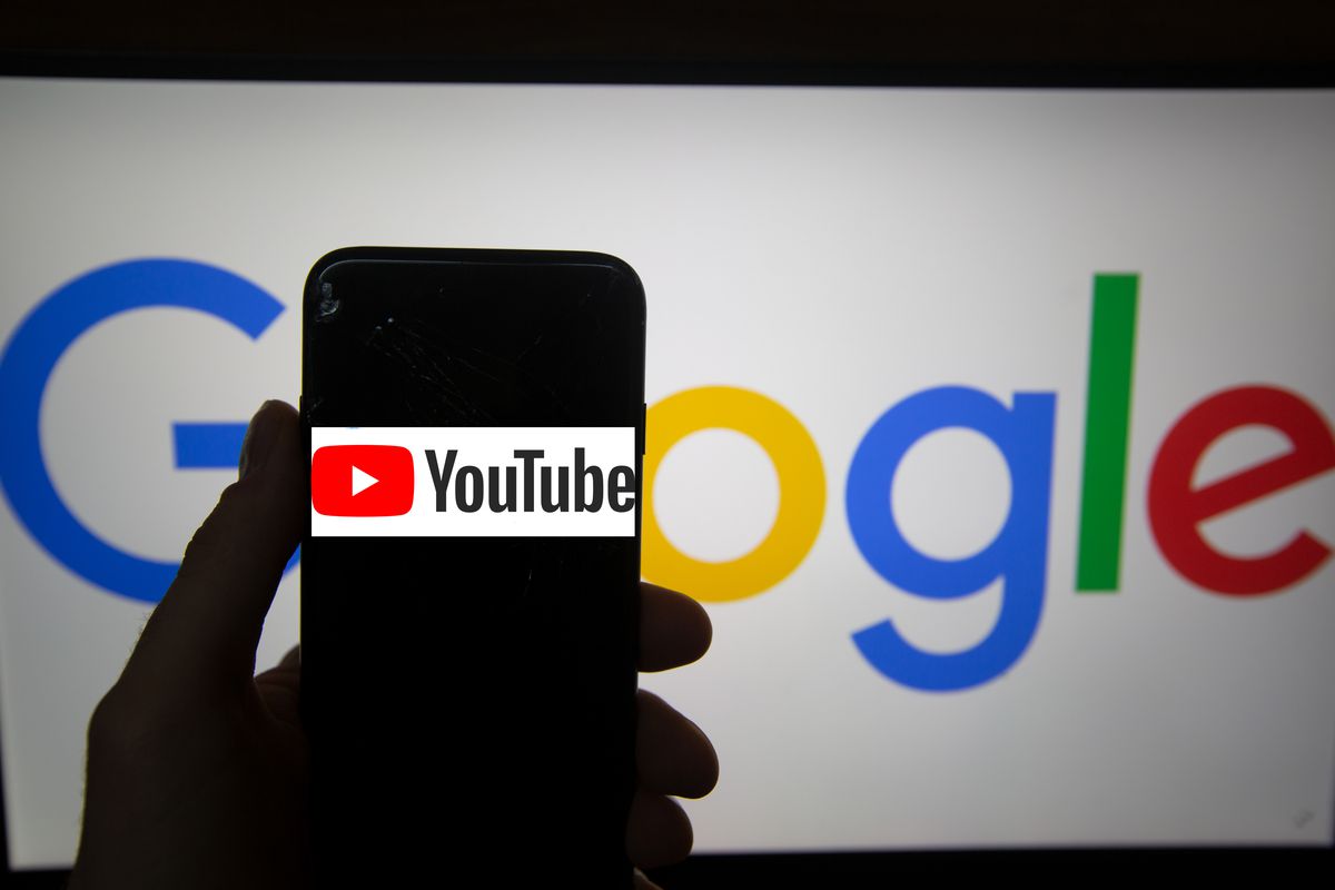 The YouTube logo on a mobile phone screen held by a hand in front of a sign reading “Google.”