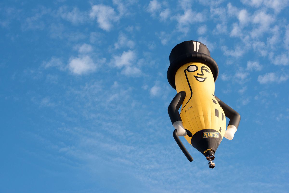 A hot-air balloon in the shape of Mr. Peanut floats across a blue sky scattered with clouds.