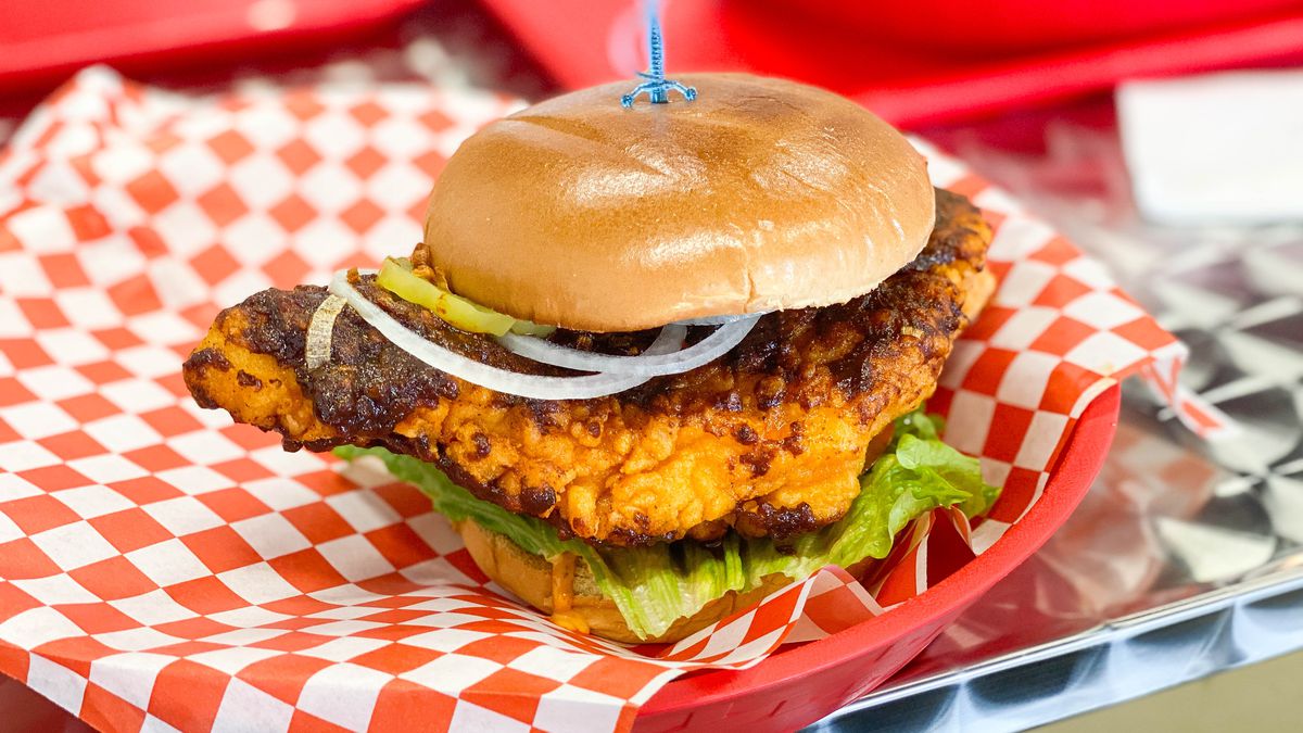A fried chicken sandwich on red checkered paper at daytime.