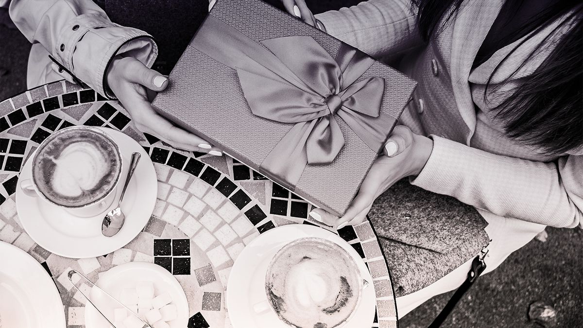 A black and white photo shows two people seated next to a round mosaic table with two lattes and their hands grasping a gift box with a large bow made from silk ribbon. 