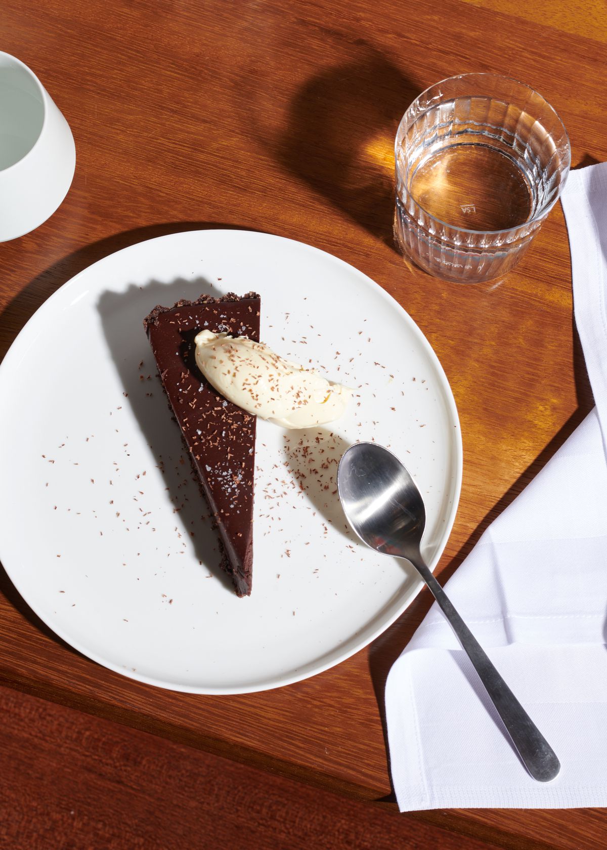 A slice of chocolate pudding pie on a white plate, with a spoon slanted aside it on a wooden table. A water glass sits to the top right.