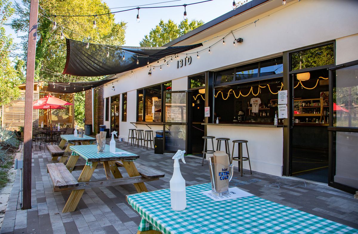 The patio at Boggs Social and Supply in West End Atlanta with three picnic tables covered in green checked plastic table cloths, spray bottles filled with sanitizing spray. String lights are hung above and the windows to the to-go bar are open