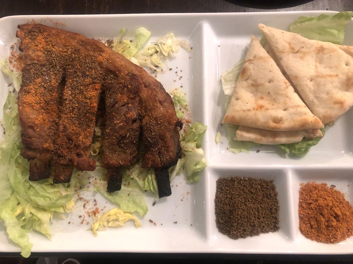 A white plate with lamb ribs, dusted with a cumin and hot pepper mix, atop some shredded lettuce. small triangles of bread and more cumin and hot pepper mix are also on the plate.
