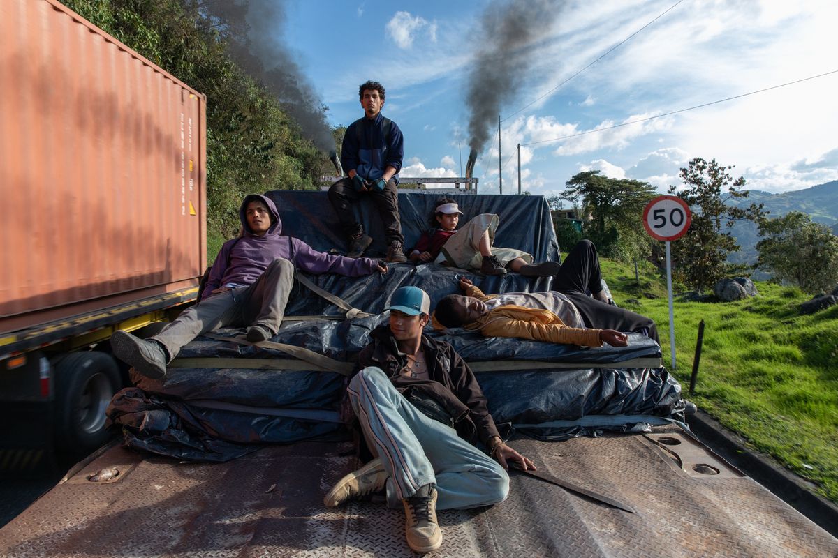 Five boys (Carlos Andrés Castañeda, Davison Florez, Brahian Acevedo, Cristian Campaña, Cristian David Duque) sit atop a mound of tarps on the bed of a train car flanked by trees, an orange storage container, and a blue sky in the background.