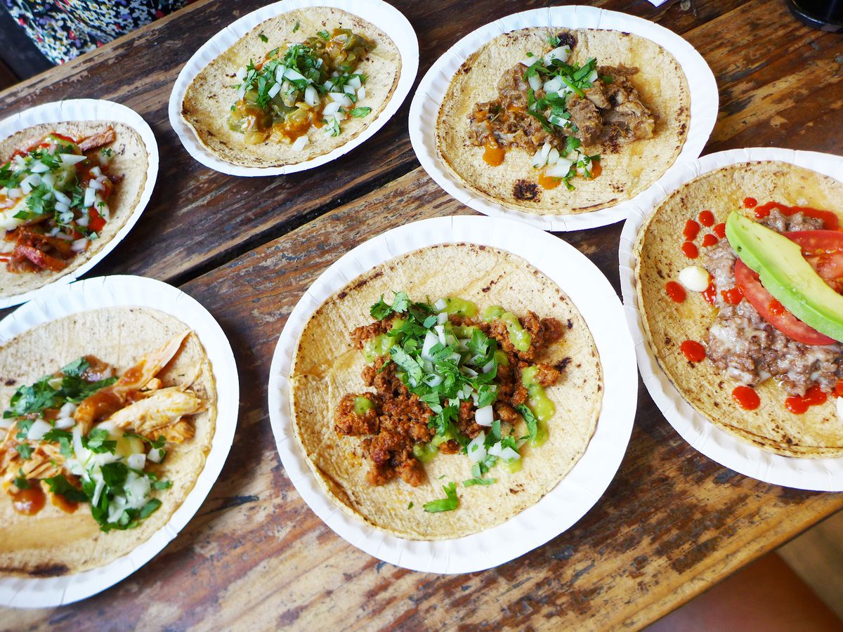 Six tacos, each on its own paper plate, including ingredients like ground beef, sliced avocado, pork chunks, and bright green cilantro.