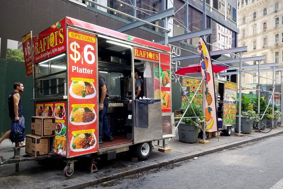 A halal cart and a smoothie cart on a midtown Manhattan sidewalk crowded with stone planters and construction scaffolding. 