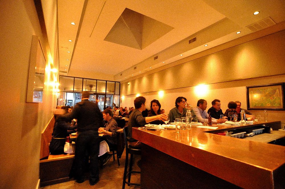 Patrons at the bar and dining room of Animal in March 2014 with orange lighting, wine glasses at the counter.