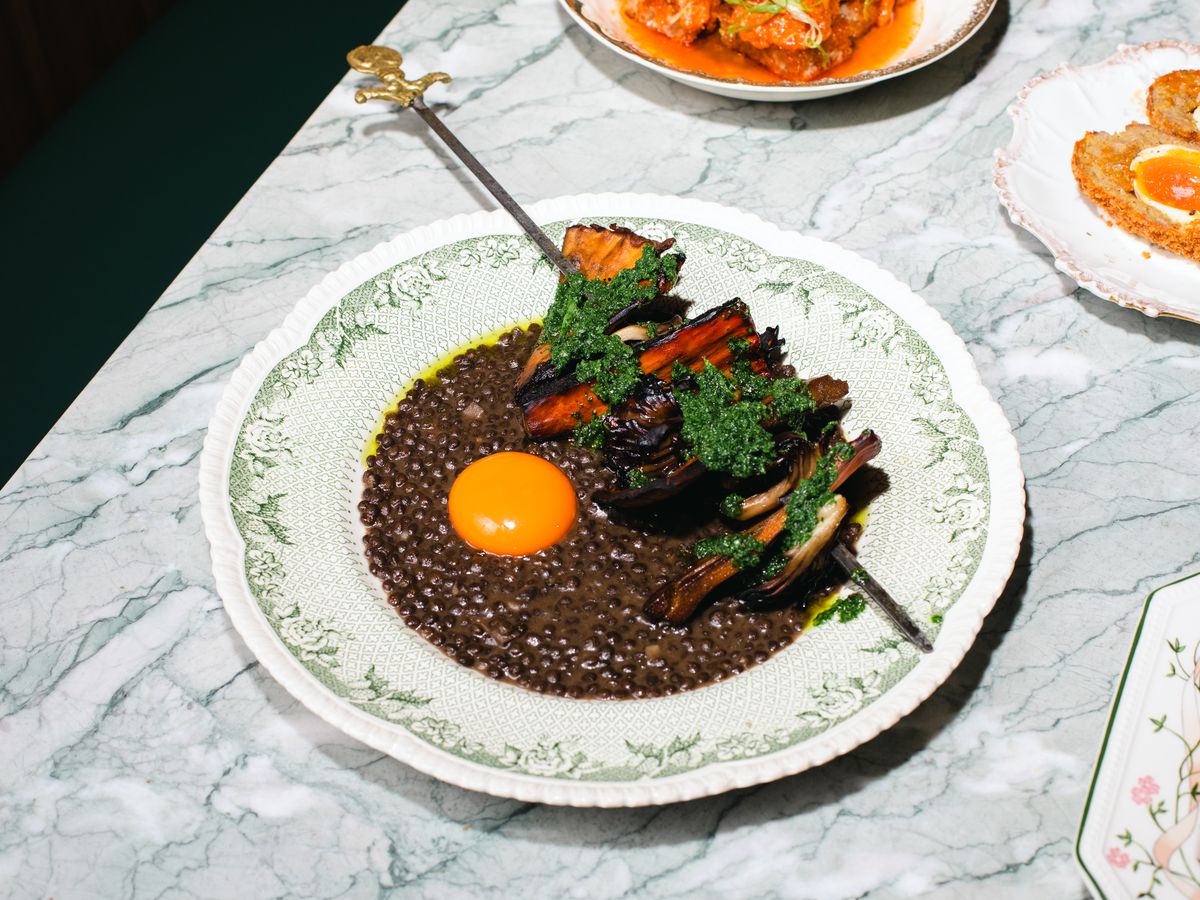 A wide plate layered with black lentils, an orange egg yolk, and a skewer of mushrooms laid over top.
