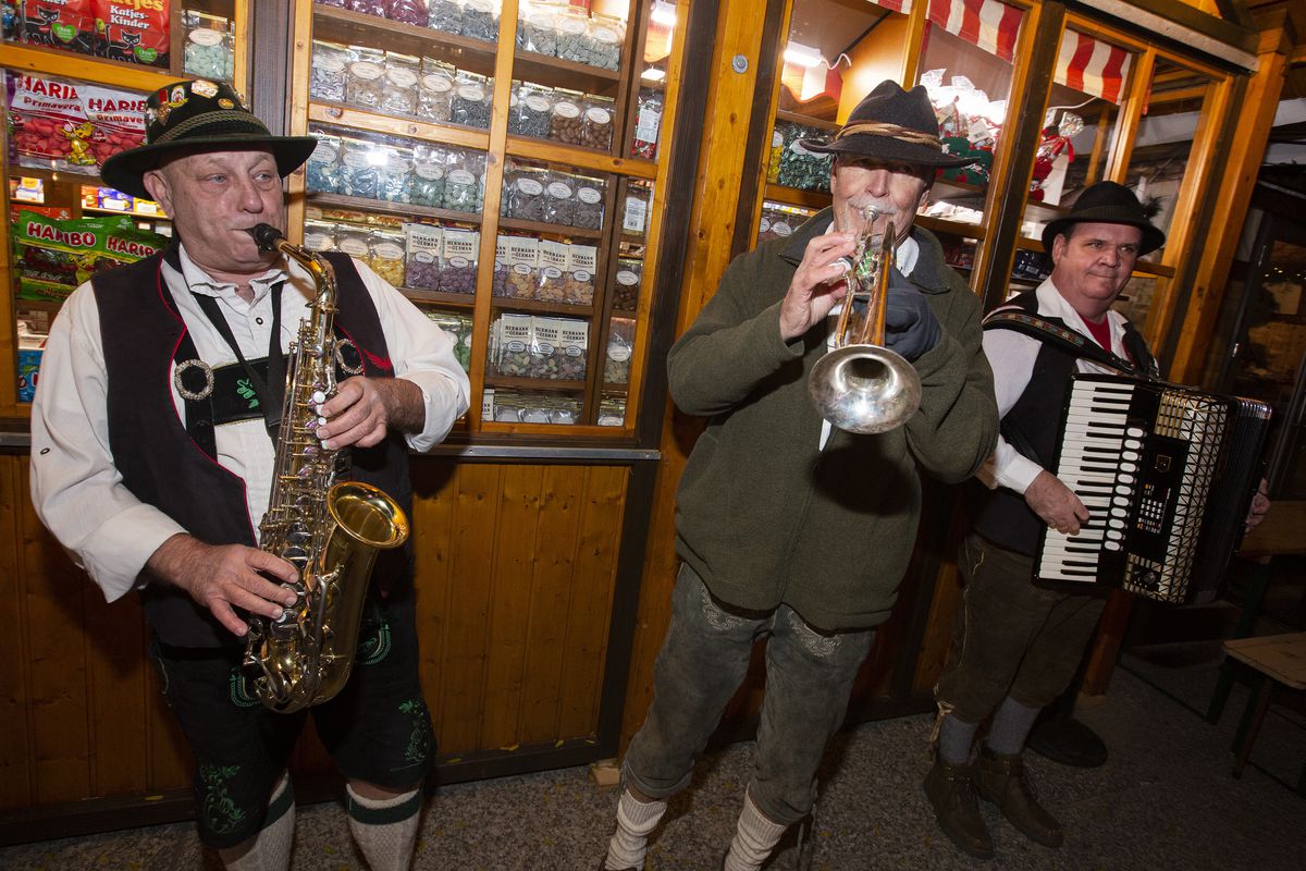 Three men wearing lederhosen play a saxophone, a trumpet, and an accordion stand outside a wooden building.