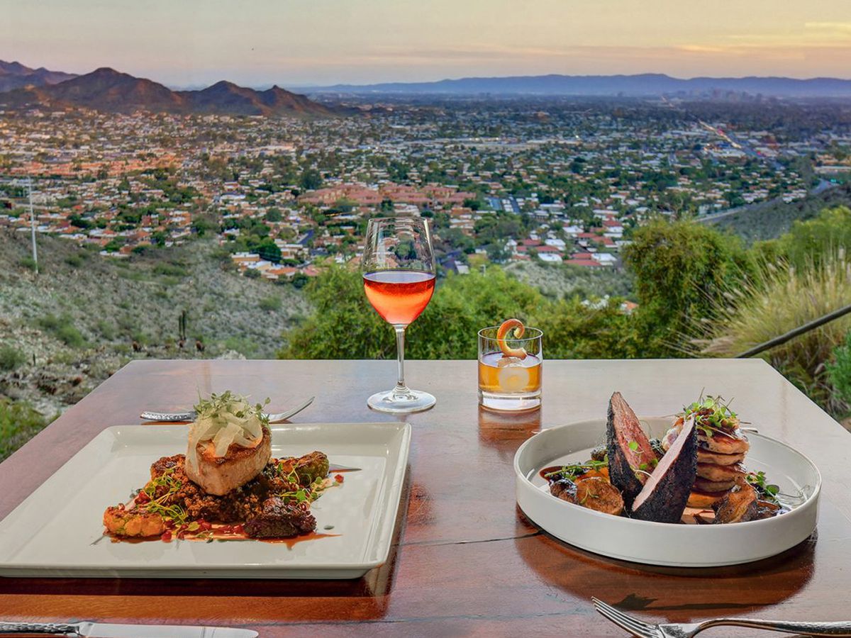 Two plates of food and a rose-colored beverage in a stemmed glass overlook a multi-colored sky and an aerial view of the city below.