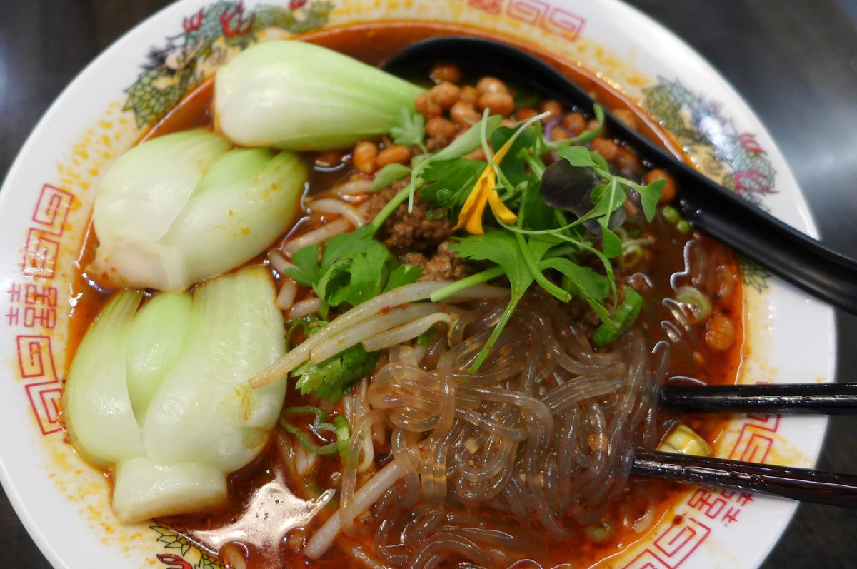 A reddish broth in a bowl with bok choy and all sorts of other stuff, including clear noodles.