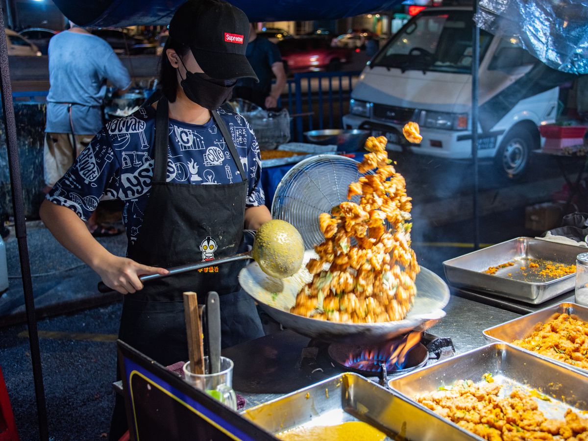 A woman in a Supreme branded hat flings egg yolk chicken into the air from her wok