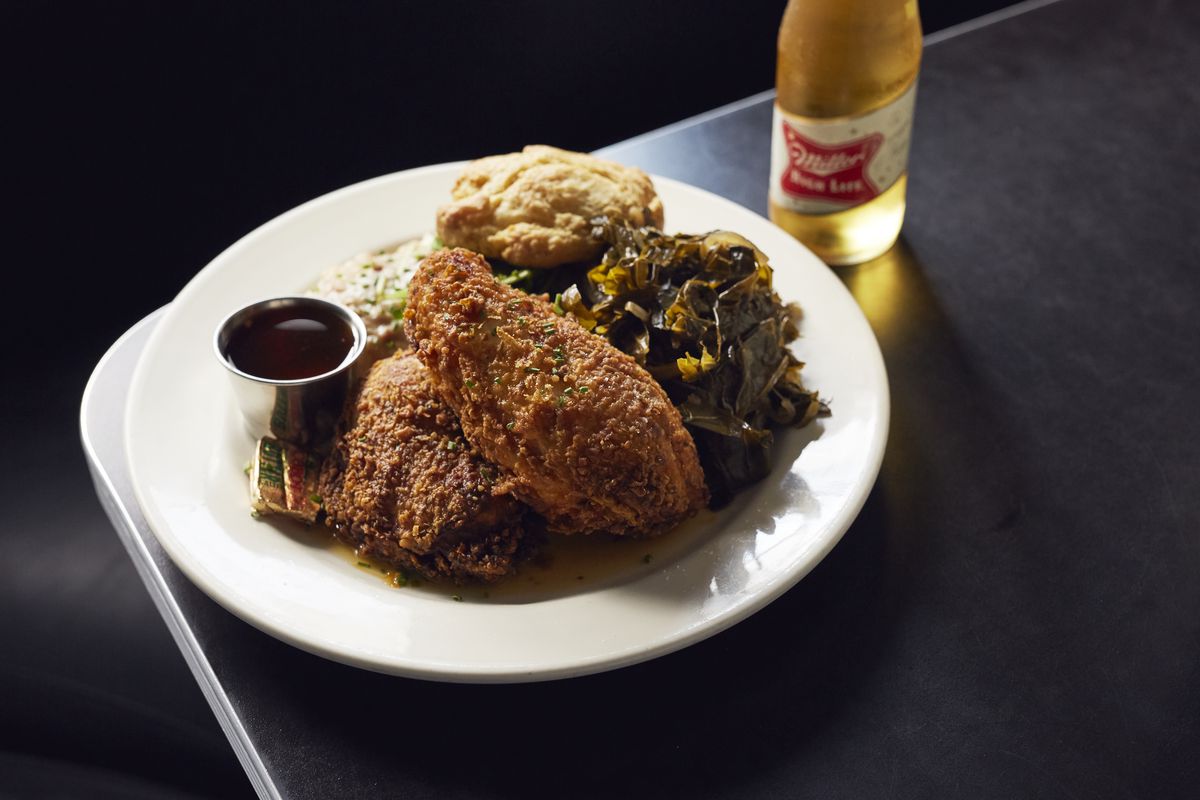 A plate of fried chicken with collard greens and a biscuit sits on a black tabletop on a black background. There’s a bottle of Miller High Life to the side of the plate.