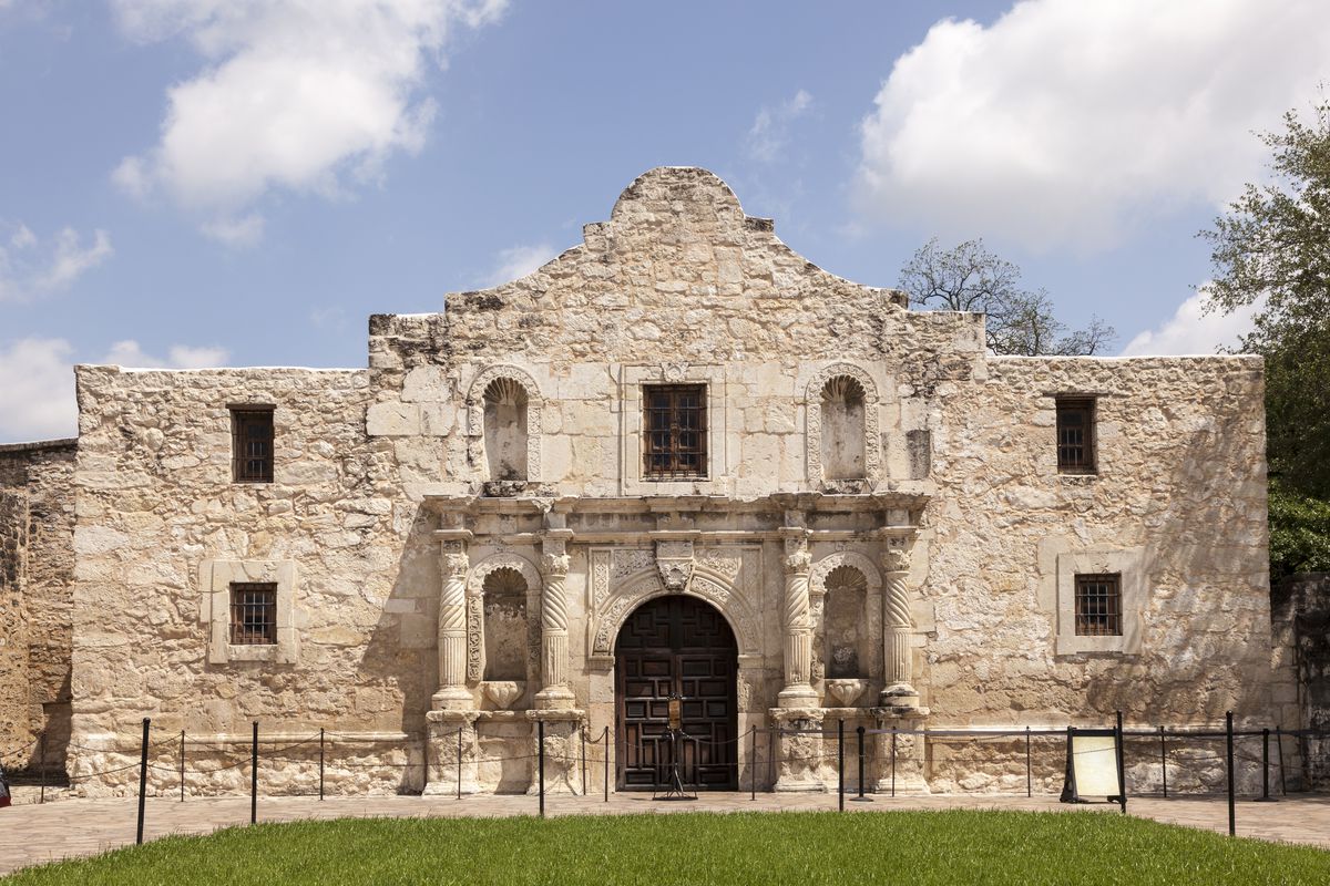 The Mission San Antonio de Valero building in Texas (known as the Alamo). The building’s facade is stone. The door is large, wooden, and arched. There are columns on both side of the door. The windows are small and have bars.