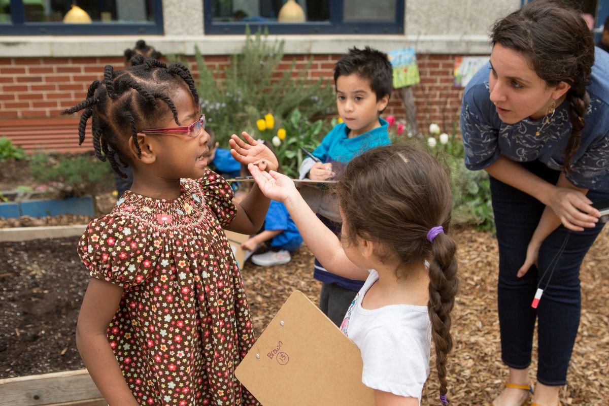 A preschool girl stands outside holding an insect on her finger while a classmate and a teacher look at it. 