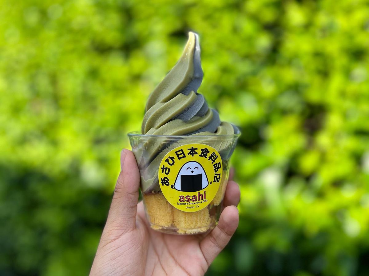 A hand holding up a clear plastic container of green soft serve in front of a green bush. 