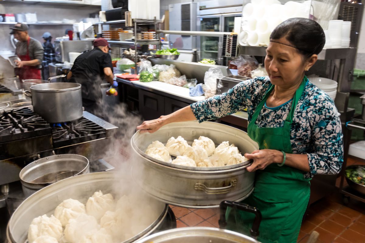 A woman lifts a giant steamer filled with soft, white steamed buns, onto a rack with even more buns inside a busy kitchen