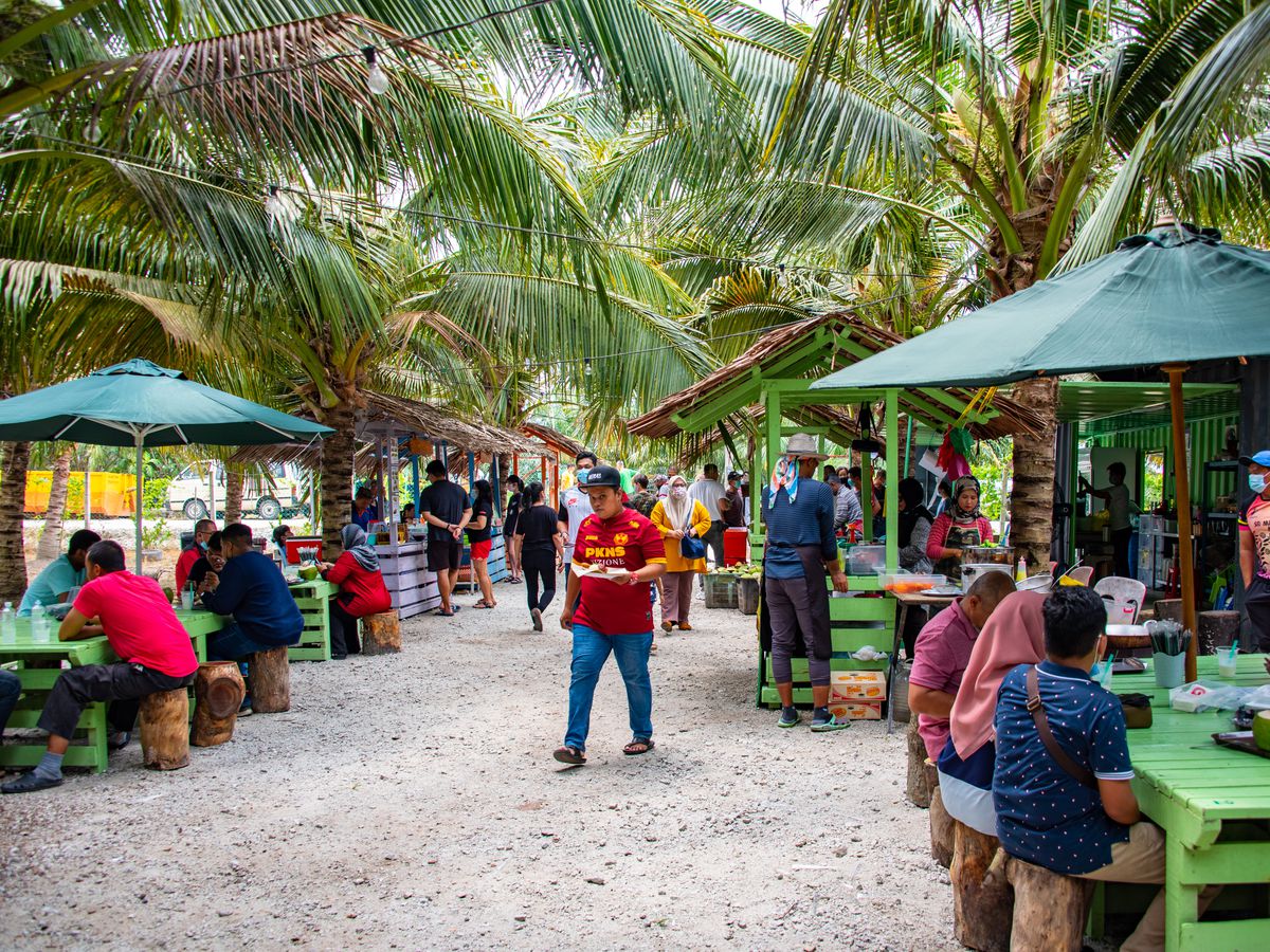 Diners eat at outdoor wooden tables and purchase food from vendors in the background, beneath rows of coconut palms