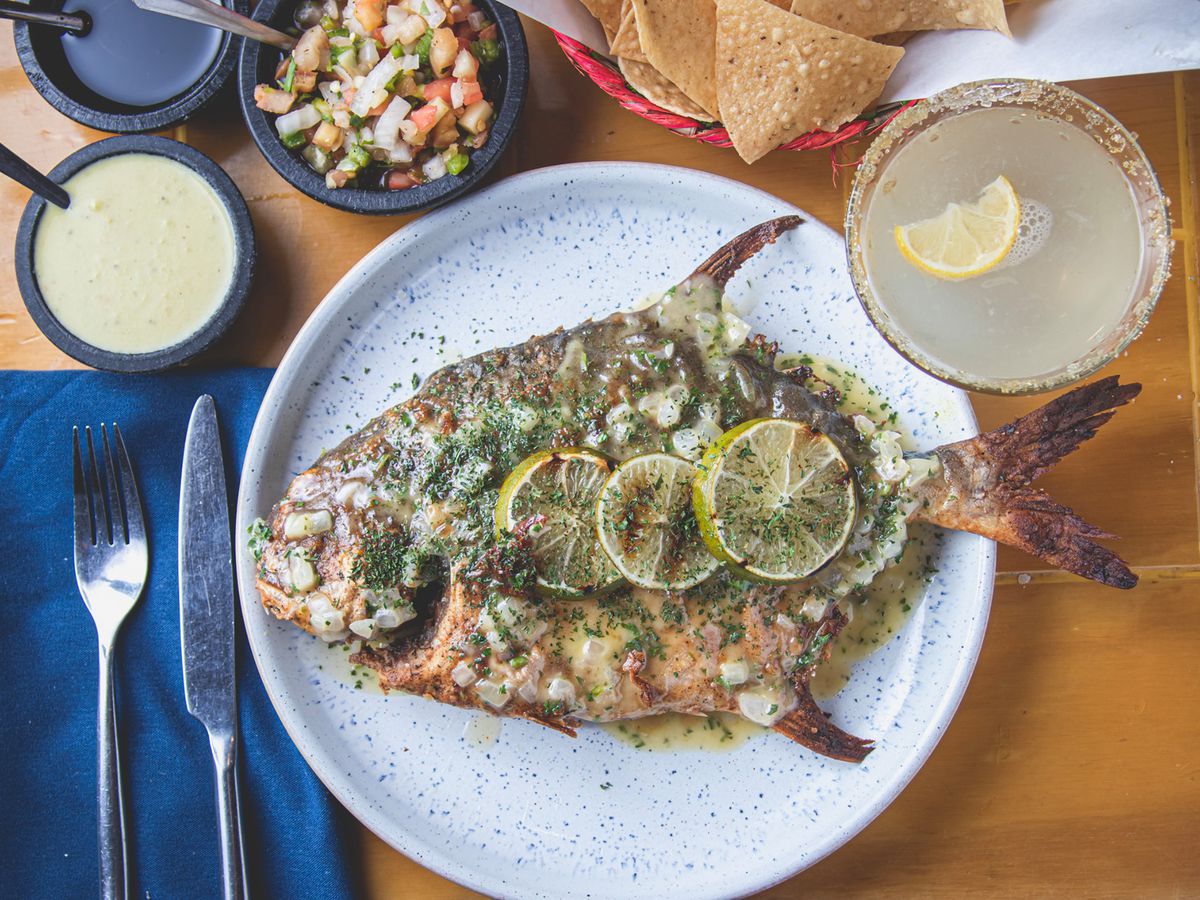 a overhead shot of a whole roasted pompano fish topped with lime slices and flanked by a margarita and various sauces