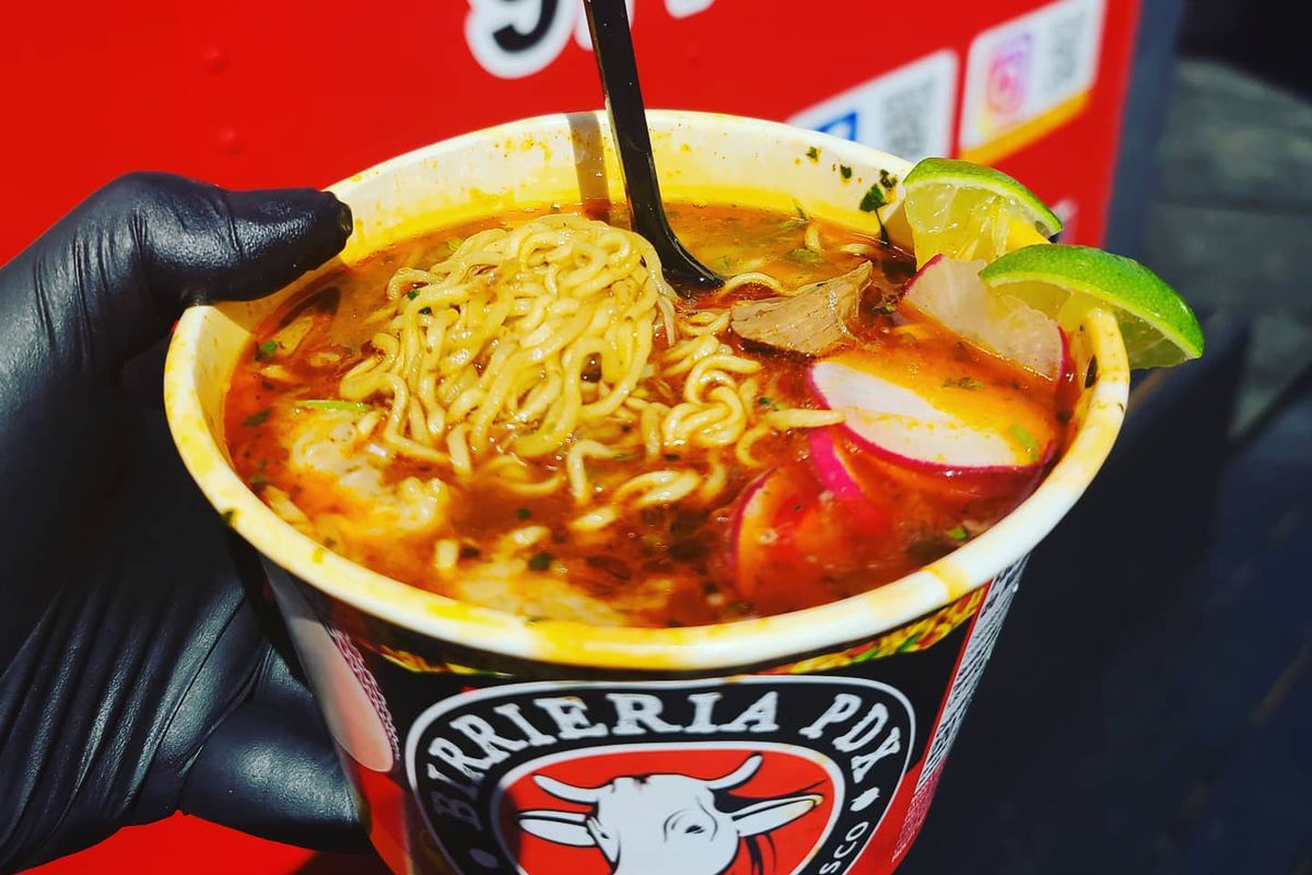 A gloved hand holds a bowl of birria with ramen noodles, radishes, and slices of lime