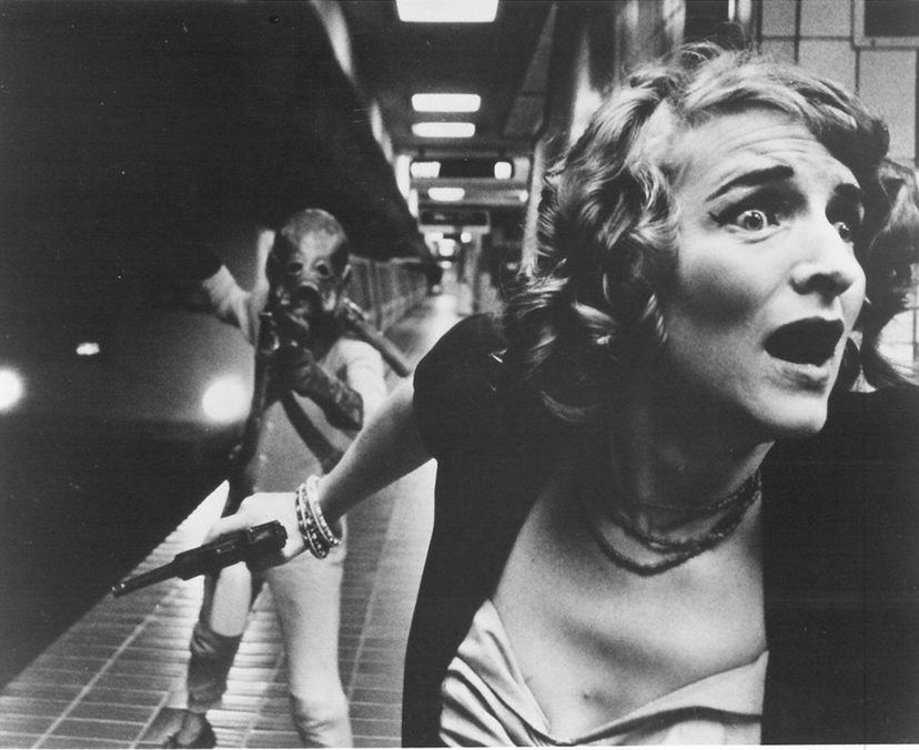A black and white image of a woman holding a gun running away from an alien monster with a huge head on a BART platform.