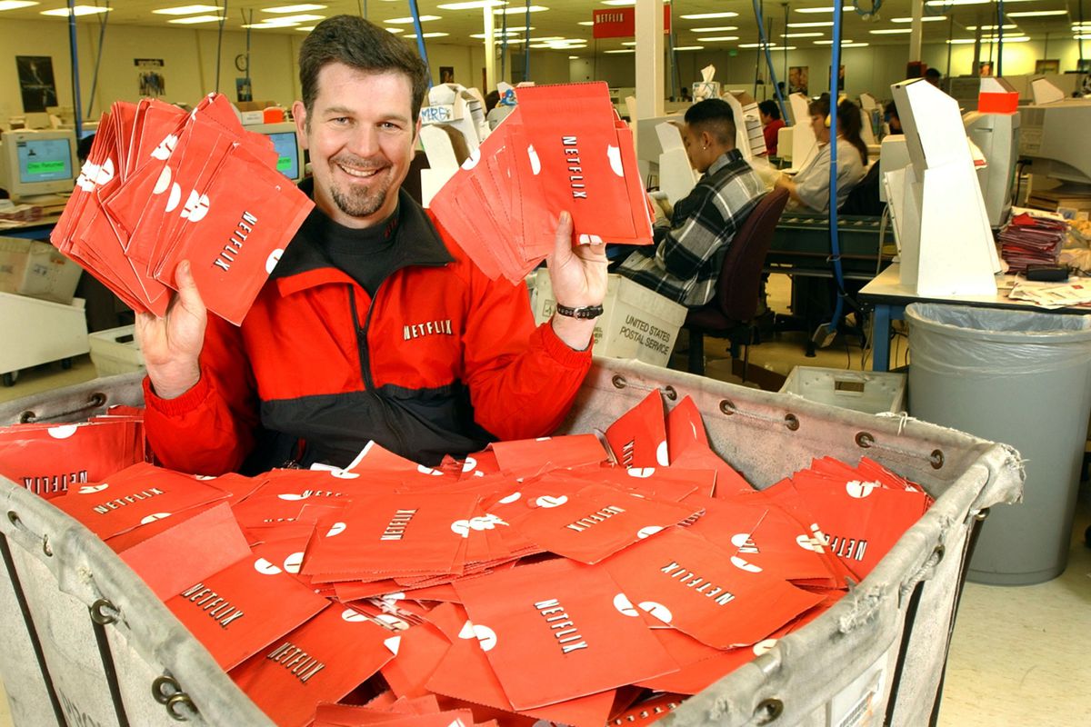 Netflix founder Reed Hastings sitting in a mail crate full of red DVD envelopes.