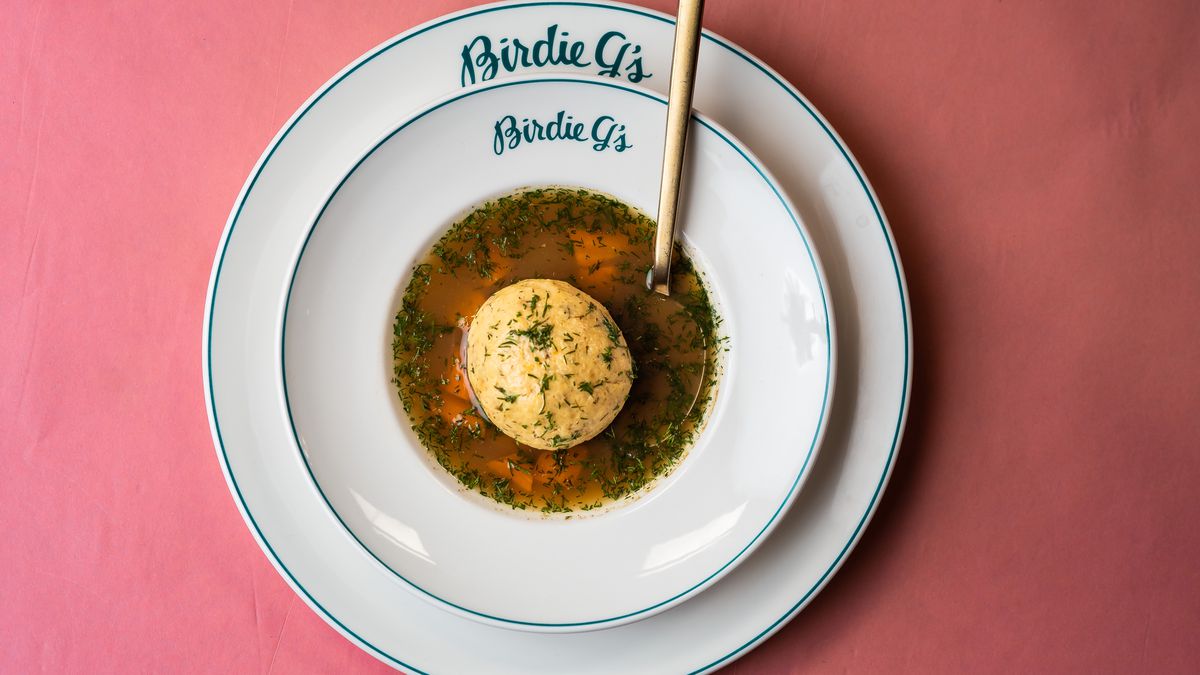 White bowl filled with matzo ball soup atop a white plate inscribed with Birdie G’s.