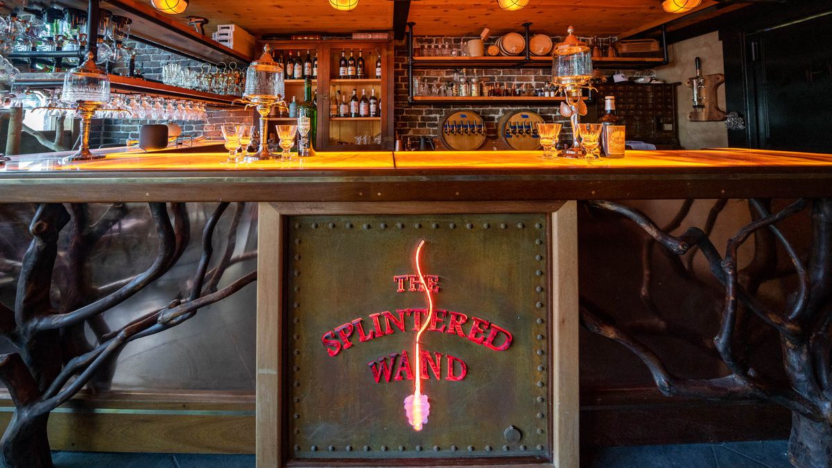 A bar lit with soft orange overhead lamps, decorated with a sign that says “The Splintered Wand” in glowing red letters and branches of a plant snaking below.