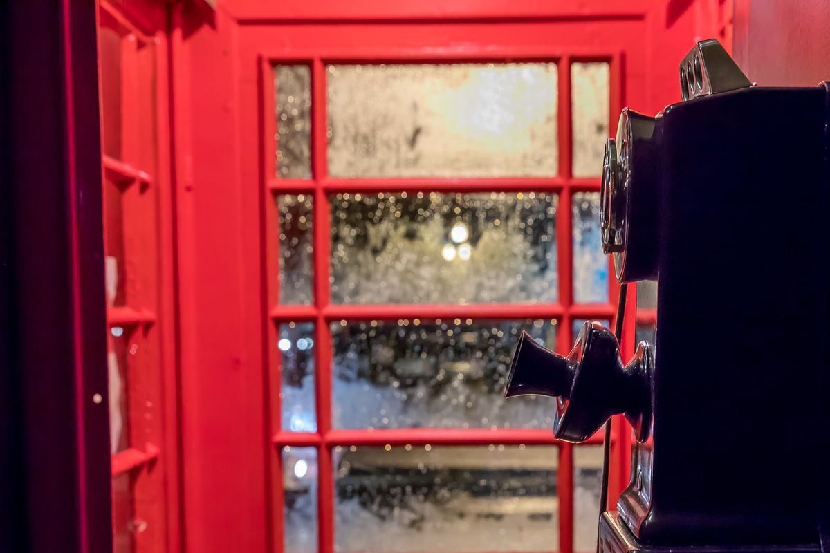 An old fashioned black telephone sits inside an red antique British phone booth as rain drops trickle down the closed glass door