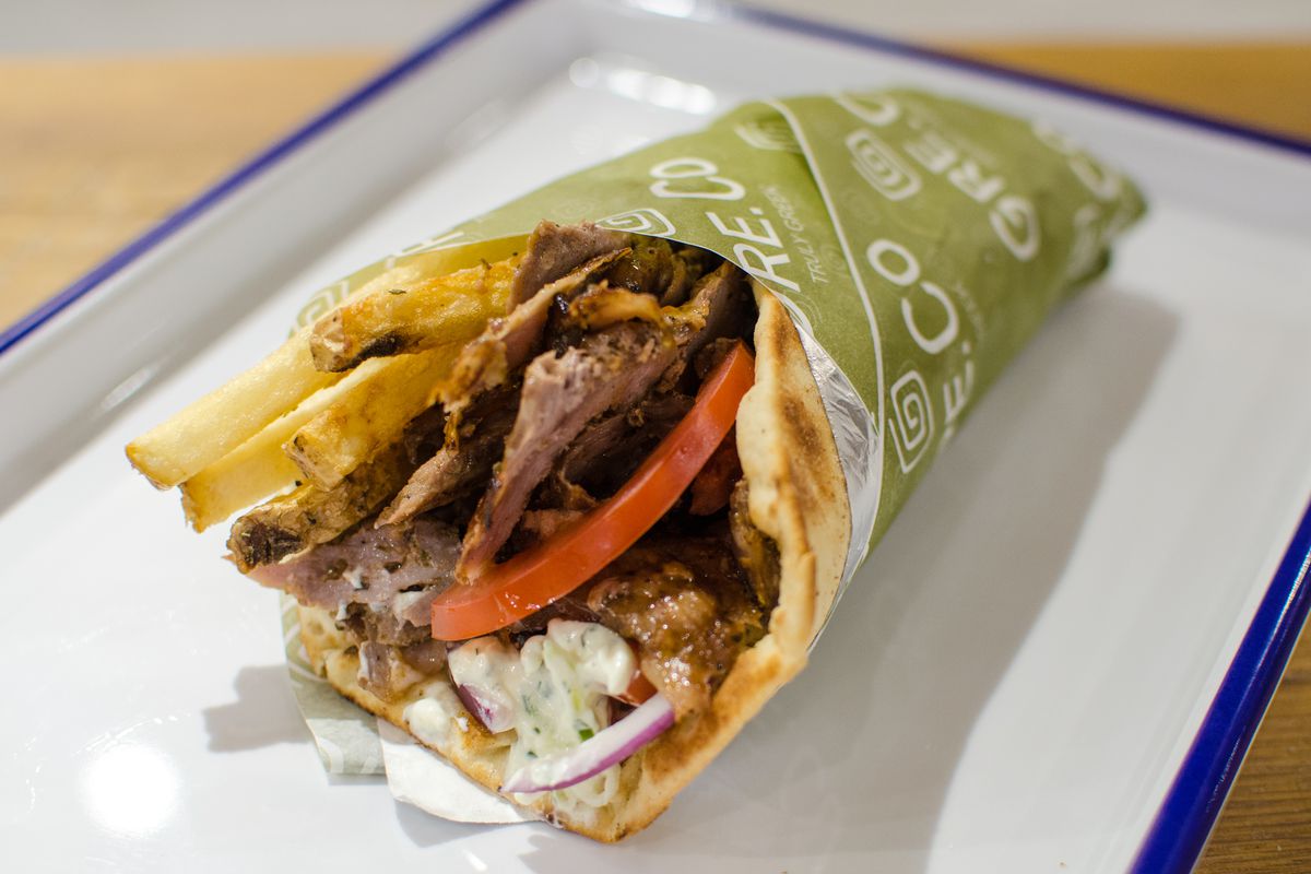 A meat gyro stuffed with fries, red onion, and tzatziki is wrapped in green paper and sits on a white tray on a wooden table.