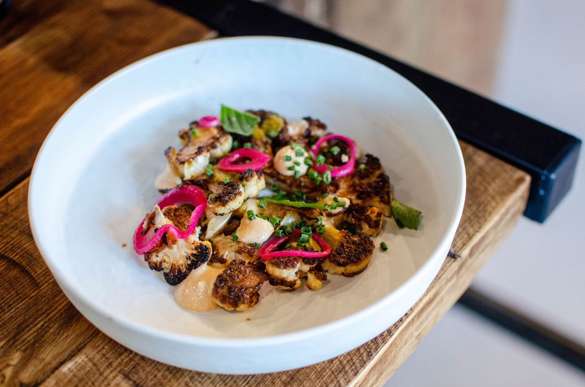 Charred bits of cauliflower sit on a white plate with dollops of whiteish-red sauce, fresh basil, chives, and pink rings of pickled onion.