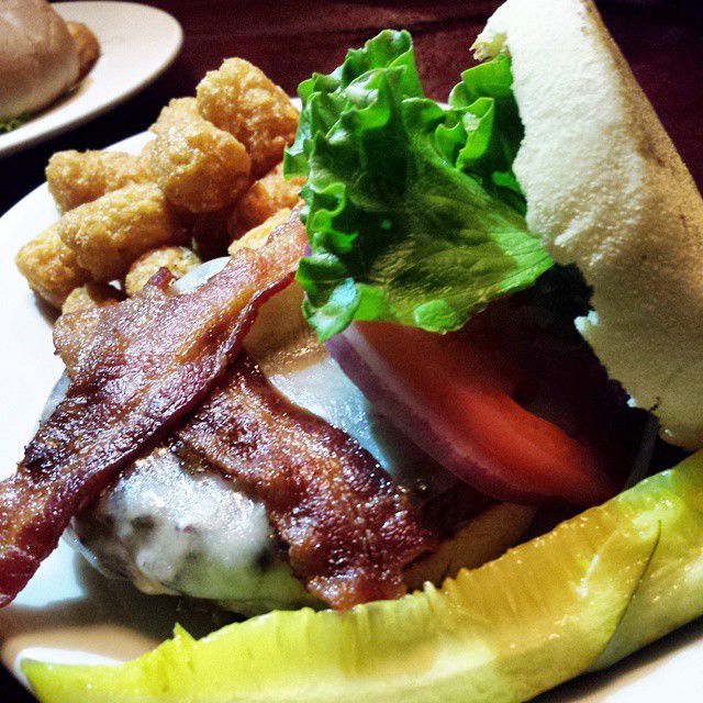 Burger on an English muffin, topped with bacon, cheese, lettuce, and tomato. A side of tater tots is visible in the background.