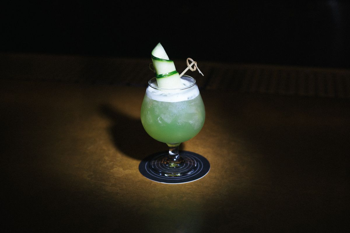 A brandy glass is filled with green liquid, with a cucumber slice rolled inside and held together with a toothpick. 