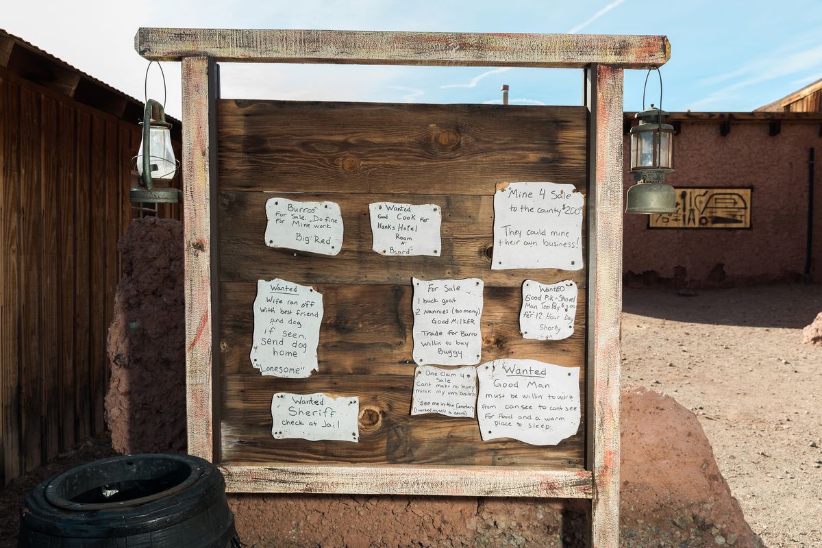A large wooden sign board with two lanterns hanging. There are handwritten signs attached to the board. The board is outside in a desert setting.