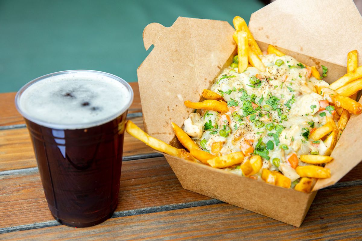 A side angle shot of a picnic table holding a cardboard box of cheesy fries and a dark beer in a plastic, clear cup.